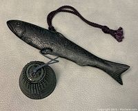 Top angled view of metal fish with attached bell and purple hanging cord on white background.