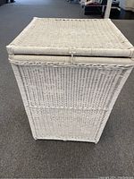 Front view of large white wicker basket with closed hinged lid on a carpeted floor.