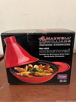 Front view of the Maxwell & Williams Microstoven Tanjine pot box showing the red and black non-stick pot with food inside and brand labeling.