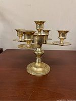 Front angled view of a solid brass 5-taper candle holder candelabra on a wooden surface, showing central elevated taper holder with four arms and drip plates.