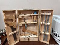 Front view of the open wooden box containing the toy tool set, including a saw, hammer, wrench, screwdriver, and other wooden and metal tools arranged on shelves and hooks inside the box doors.
