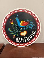 Front view of round vintage folk art sign with blue bird, floral artwork, heart, and Wilkom script on black background with red scalloped border
