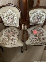 Photo of two vintage wooden chairs with cream-colored floral needlepoint upholstery and carved wooden frames showing detailed ornate design and fabric pattern