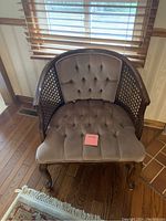 Beige tufted velvet accent chair with lattice wood sides and carved wooden legs, placed on a wooden floor in front of a window with blinds.