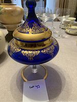 Photo of an ornate blue covered dish with gold filigree detailing on a pedestal base, on a table with other glassware.