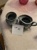 Cream and sugar set displayed on a beige cloth surface with a note showing lot number 39. The ceramic items have blue and earthy glaze.