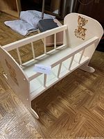Wooden doll cradle shown from an angle, displaying side railings and deer artwork on headboard and footboard, resting on a hardwood floor.