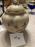 Light beige Nippon porcelain covered jar with pink floral motifs and gold rim on lid, displayed on a fabric surface with other items in the background.