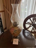 Antique hurricane lamp with floral glass reservoir and clear glass chimney on a metal base, photographed indoors near window.