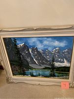 Framed oil painting depicting a scenic mountain lake landscape with snow-capped peaks, pine trees, and reflective water.