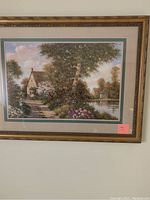 Framed landscape picture showing a house, garden, and water body with trees and flowers, in a gold ornamental frame.