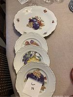 Four porcelain plates arranged with the large platter at the back and three smaller plates stacked in front. Each plate has a scalloped edge and painted fruit design.