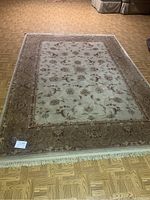 Photo of full view of large rectangular woven rug laid out on parquet floor showing detailed floral and border pattern in beige and muted reds with fringe on edge.