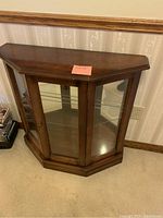 Front angle view of a small wooden curio cabinet with a glass door and shelf, showing the hexagonal shaped top and clear glass sides.