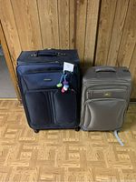 Photo showing two soft-sided suitcases against a paneled wood wall on a patterned wood floor, one dark blue and larger, the other gray and smaller. Both have front zippered pockets and wheels.