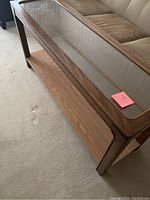 Wooden console table with glass inset top and a lower wooden shelf on carpeted floor next to a sofa.