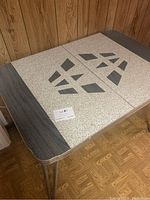 Tabletop close-up showing the speckled beige laminate with dark gray geometric design and gray faux wood grain edges, metal legs partially visible.