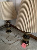 Photo showing both brass lamps with cream pleated lampshades placed on carpeted floor near wall.