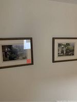 Two framed garden pictures hung on a beige wall. Both have brown frames and glass covering. The left picture shows a garden pathway and the right shows a garden with a white house.