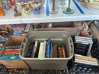 Three plastic tubs side by side filled with books, plus surrounding household items on table