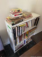 Photo of white bookcase filled with multiple art and poetry books, including some stacked on top. Shelf and books show signs of dust and need cleaning.