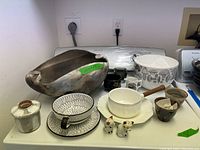 Mixed ceramics and kitchenware arranged on shelf including pit-fired pot, sugar pot, teacup, saucer, leaf-patterned bowl, enamel saucepan, cat shakers, small jars, and more