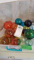 Collection of various colored glass orbs in a plastic container showing orange, green, blue, and multicolor orbs with sizes ranging from 2 to 4 inches.