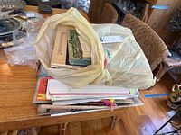 Open plastic bag with assorted vintage magazines, sheet music, and art prints stacked on a wooden table.