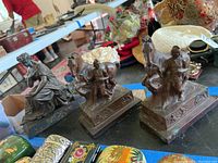 Three metal figurines on a table with a blue tape line visible, showing two Roman soldier and horse pairs and one seated female figure.