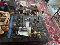 Photo showing two brass-colored candlesticks, black metal candlabra with four candle holders, and three long-handled branding irons with letters D, H, and J on the tips placed on a black table with worn surface and a patterned carpet in background.
