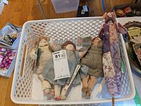 Photo showing five handmade fabric craft dolls lying in a plastic basket on a wooden floor. Dolls dressed in various vintage style clothes.