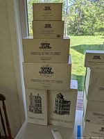 Stacked boxed items of Heritage Village Collection, showing Christmas in the City series boxes and others with clear labeling and images of buildings.