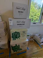Four original boxes of Heritage Village Collection ceramic buildings stacked. Two Snow Village and two Dickens Village series buildings shown.