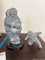 Two carved Inuit soapstone figures placed side by side on a wooden furniture surface, the larger figure mounted on a black wood round stand with scratches.