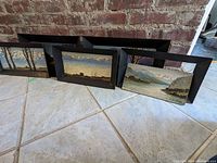View of all five framed landscape paintings leaning against a brick wall showing their size and frames.