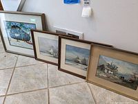 Four framed prints leaning against wall on floor. Various landscape and seascape scenes visible. Frames show minor to moderate wear.