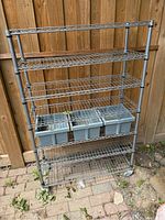 Full view of the metal shelving unit outside against a wooden fence showing seven wire shelves and three gray plastic bins on one shelf.