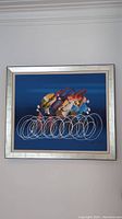 Front view of abstract painting showing bicycle race theme with overlapping circular wheels on dark blue background, framed in silver blue wood frame.