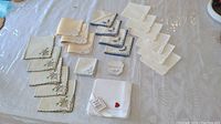 Overview of multiple hand-stitched linens arranged on a table showing cream and white items with some floral embroidery and scalloped edges.