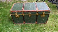 Front view of green antique steamer trunk on grass, showing metal latches and locks with brown trim.