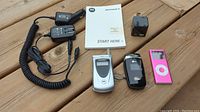 All items on wooden surface including Motorola flip phone, Sprint flip phone, pink iPod Nano 4GB, two car chargers, and Motorola charger and manual.