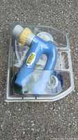 Blue cordless power scrubber with a yellow bristle brush attached, contained in molded plastic packaging with other brush attachments visible.