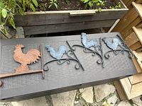 Three metal rooster wall hooks laid flat on a table, two are black matching hooks with curled designs, one is a red cast iron rooster with flat base.