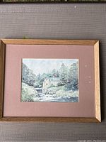 Framed painting showing a mill building by a river with trees in background, detailed landscape.