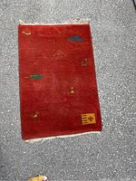 Top-down view of the rectangular handwoven wool rug on a paved surface showing the maroon base color and small colorful motifs spaced across the surface with a small fringe on the top edge.