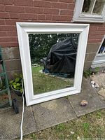 Full view of mirror leaning against brick wall outside. Shows white molding frame and reflective glass surface.