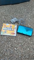 Photo showing the Nintendo 3DS aqua blue console closed, power adapter charger cable, and New Super Mario Bros 2 game case on a gravel surface