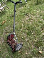 Side view of manual push lawnmower on grass showing black handle grips and curved steel handle.