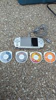 Sony PSP console with screen and four UMD game discs laid out below with charger cable in background on pavement surface.