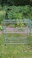 Full view of the steel wire shelving unit standing on grass outdoors with green leafy plants behind it.